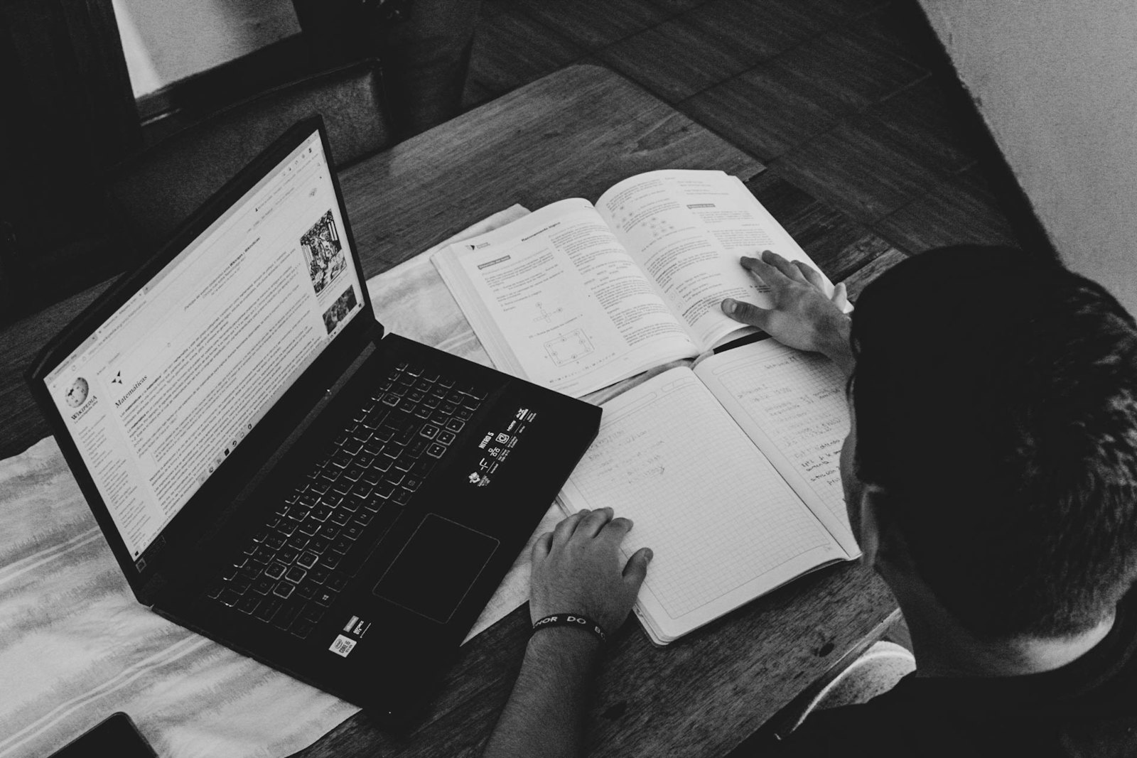 A student at a kitchen table with a laptop and an open textbook, seen from above and behind. Black and white.