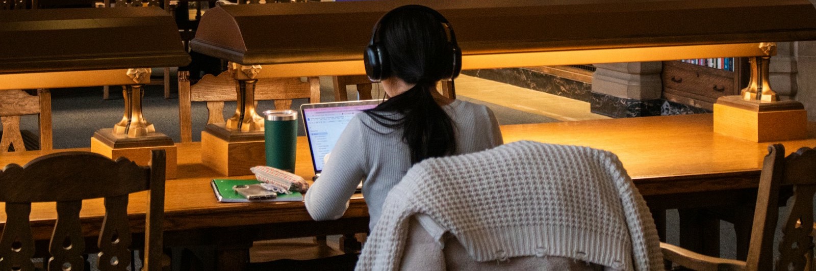 A student sits at a long wooden table in a library reading room with a laptop, seen from behind.