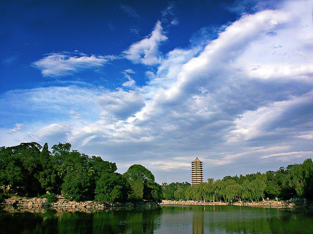 A pagoda standing above willows on the far bank of a lake, under a bright sky.